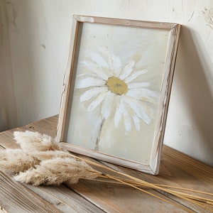 May include: A framed painting of a white daisy with a yellow center on a light beige background. The frame is made of wood and has a whitewashed finish. The painting is sitting on a wooden table with a bunch of dried pampas grass.