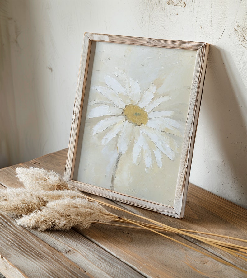 May include: A framed painting of a white daisy with a yellow center on a light beige background. The frame is made of wood and has a whitewashed finish. The painting is sitting on a wooden table with a bunch of dried pampas grass.