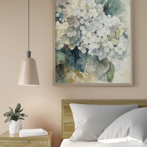 May include: A framed watercolor print of white and light blue hydrangeas with green leaves. The artwork hangs above a bed with white bedding and gray pillows. A nightstand with a lamp and a potted plant is next to the bed.