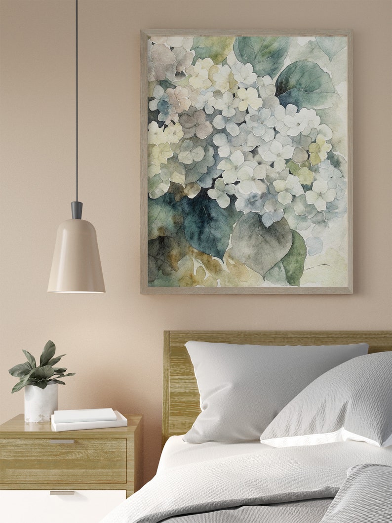 May include: A framed watercolor print of white and light blue hydrangeas with green leaves. The artwork hangs above a bed with white bedding and gray pillows. A nightstand with a lamp and a potted plant is next to the bed.