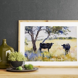 May include: Watercolour painting of two black cows standing in a field under a tree. The cows are facing different directions and the field is a light yellow colour. The tree is a dark green colour and has a thick trunk. The painting is framed in a light wood frame.