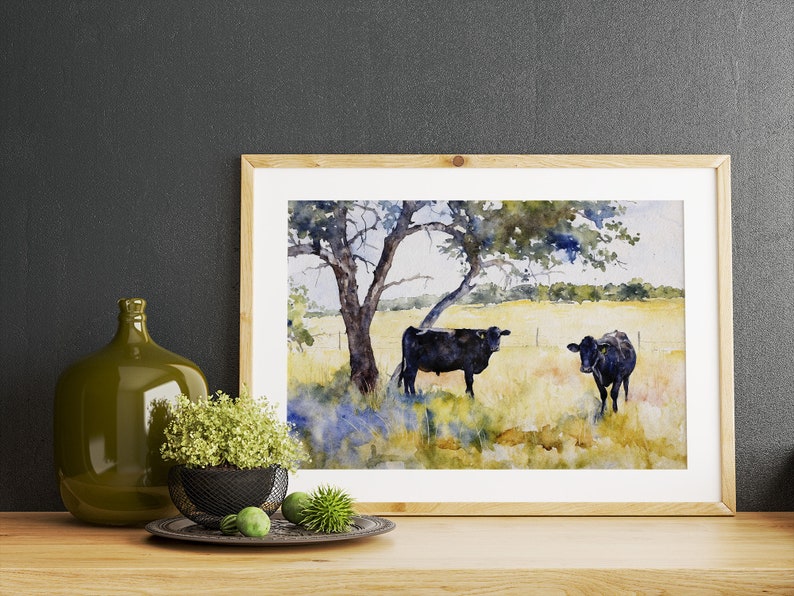 May include: Watercolour painting of two black cows standing in a field under a tree. The cows are facing different directions and the field is a light yellow colour. The tree is a dark green colour and has a thick trunk. The painting is framed in a light wood frame.