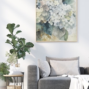 May include: A framed watercolor print of white and blue hydrangea flowers. The artwork has a light wooden frame and is displayed on a white wall above a gray sofa. The painting features soft colors and a botanical theme.