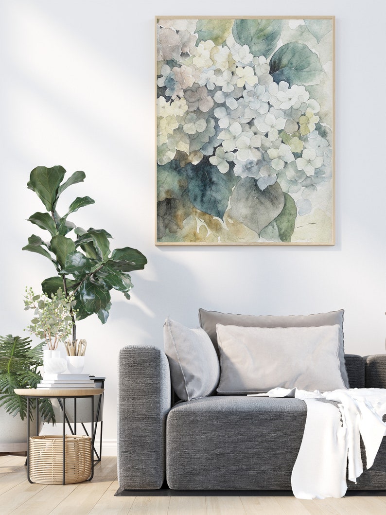 May include: A framed watercolor print of white and blue hydrangea flowers. The artwork has a light wooden frame and is displayed on a white wall above a gray sofa. The painting features soft colors and a botanical theme.