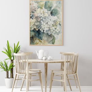 May include: A framed watercolor print of hydrangeas in shades of white, blue, and green. The artwork is displayed above a round wooden table set with a teapot, cups, and chairs. A potted plant is on the left.