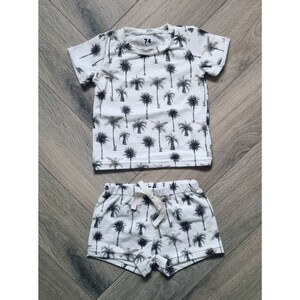 Op de afbeelding: Een witte twee-delige outfit voor een baby of peuter. Het bovenstuk is een T-shirt met korte mouwen met een palmprint. De broek is wit met een palmprint en heeft een trekkoord.