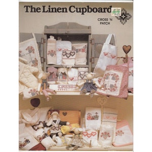 May include: A collection of handmade linen items, including towels, oven mitts, and aprons, are displayed in a rustic wooden cabinet. The items feature cross-stitch floral and heart designs in red, pink, and white. The text "The Linen Cupboard" is at the top.