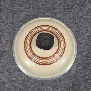 May include: A vintage Club brand pot lid. The lid is cream-coloured with concentric brown rings and a dark brown handle. The handle has the word "Club" embossed on it. The rim is silver.