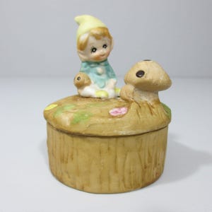 May include: A ceramic trinket box with a mushroom and a small elf figurine on top. The box is painted to look like a tree trunk and has a green and brown base.