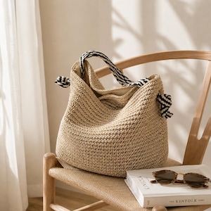 May include: A beige woven tote bag with a black and white striped handle and accents. The bag is on a wooden chair with a book and sunglasses.