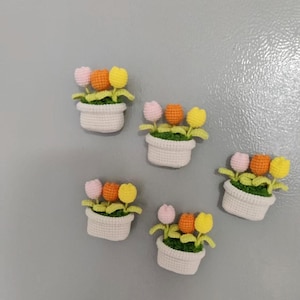Könnte beinhalten: Fünf gehäkelte Tulpen-Blumentopf-Magnete. Jeder Topf enthält drei Tulpen in Rosa, Orange und Gelb mit grünen Blättern. Die Töpfe sind weiß und haben eine gewebte Textur. Die Magnete sind auf einer grauen Oberfläche angeordnet.