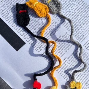 May include: Three crocheted cat bookmarks in black, yellow, and gray. Each bookmark has a red, yellow, or gray bow tie. The bookmarks are on a page of a book.