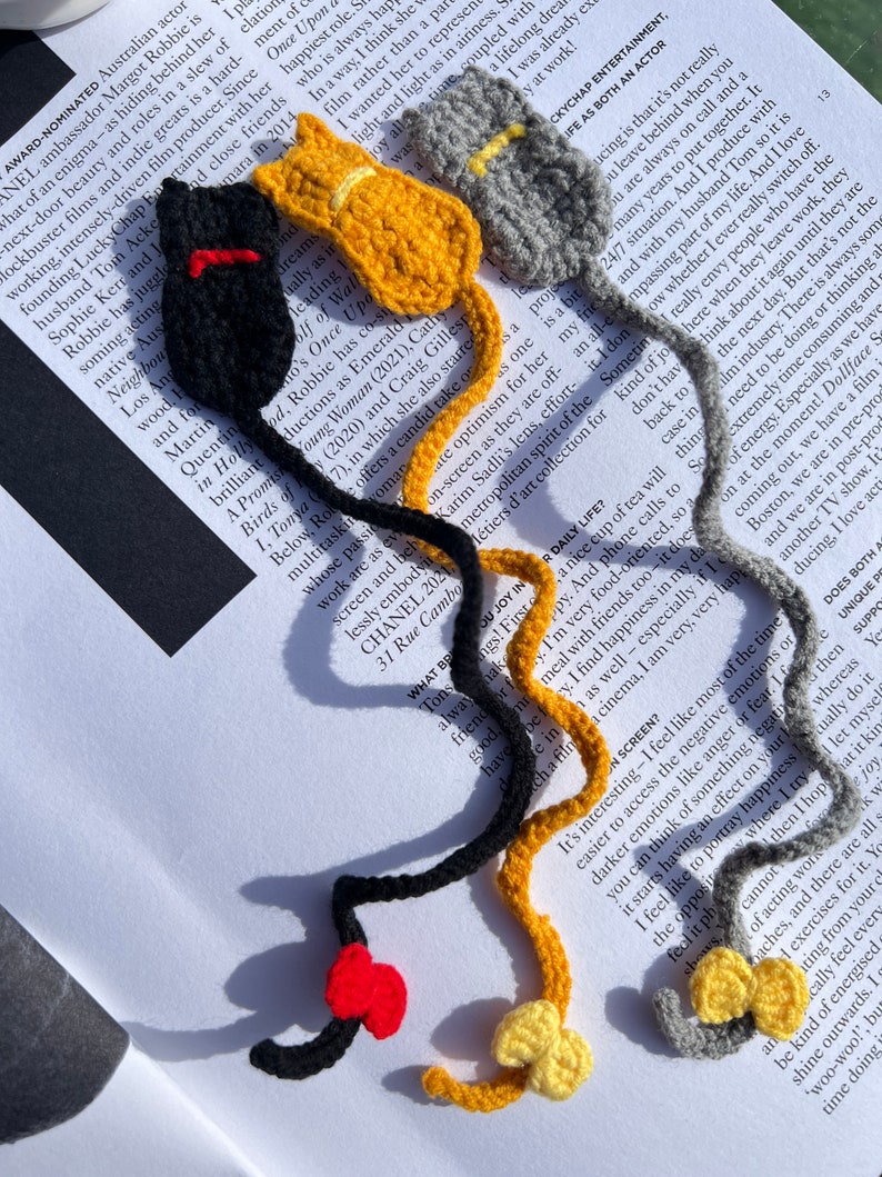 May include: Three crocheted cat bookmarks in black, yellow, and gray. Each bookmark has a red, yellow, or gray bow tie. The bookmarks are on a page of a book.