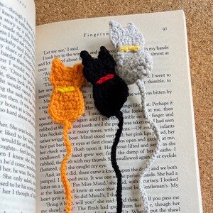 May include: Three crocheted cat bookmarks in orange, black, and gray. The bookmarks have yellow yarn accents. The bookmarks are placed on an open book with text.