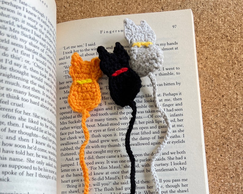 May include: Three crocheted cat bookmarks in orange, black, and gray. The bookmarks have yellow yarn accents. The bookmarks are placed on an open book with text.