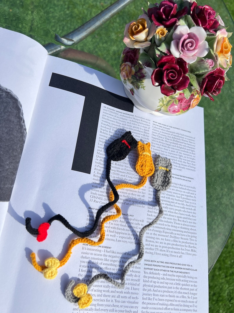 May include: A close-up of a magazine page with text about acting and producing.  There are three crocheted cat bookmarks in black, yellow, and gray.