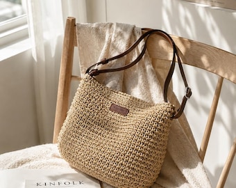 May include: A woven beige shoulder bag with brown leather straps and a small rectangular label. The bag is sitting on a wooden chair with a cream-colored blanket and a magazine with the word "KINFOLK" on the cover.