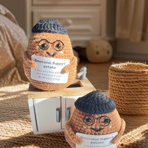 May include: Two crocheted potato figures, each wearing a black cap and round glasses. They hold signs that read "Emotional Support Potato" with an inspirational message. The potatoes are brown and sit on a miniature wooden cabinet.