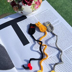 May include: Four crocheted cat bookmarks in red, black, yellow, and gray. The bookmarks are shaped like cats with long tails and are strung together.