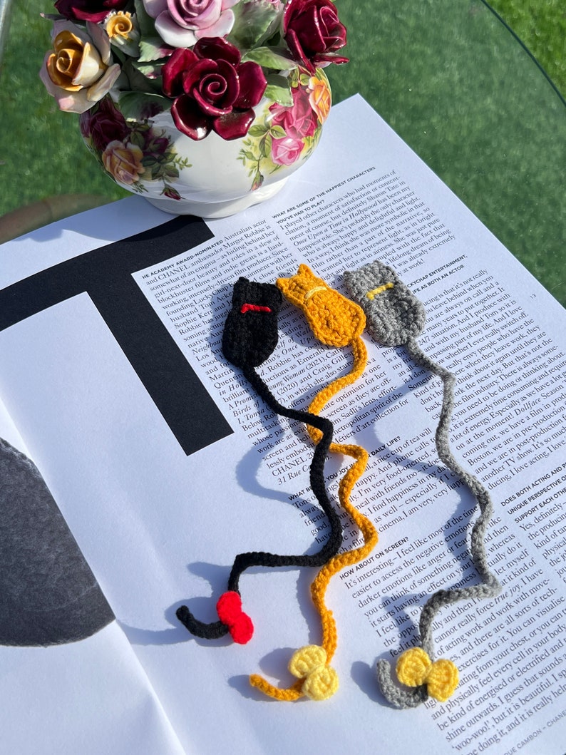May include: Four crocheted cat bookmarks in red, black, yellow, and gray. The bookmarks are shaped like cats with long tails and are strung together.