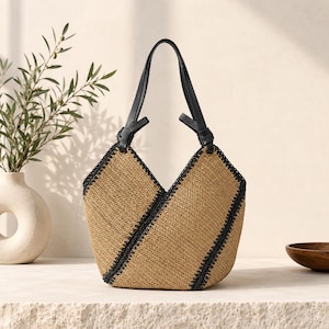 May include: A woven tote bag with a V-shaped design. The bag features a natural tan colour with black trim and handles. The handles are made of black leather and are knotted at the top. The bag is sitting on a white surface.