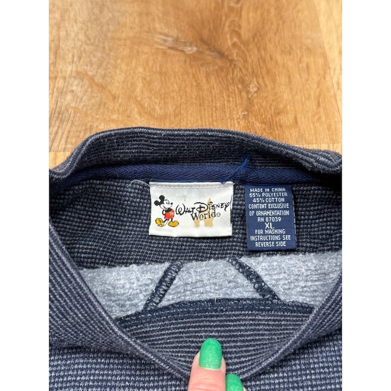 May include: Close-up of a navy blue and gray Walt Disney World sweatshirt. The tag features the Walt Disney World logo with Mickey Mouse and text that reads "MADE IN CHINA 55% POLYESTER 45% COTTON CONTENT EXCLUSIVE OF ORNAMENTATION RN 87039 XL".
