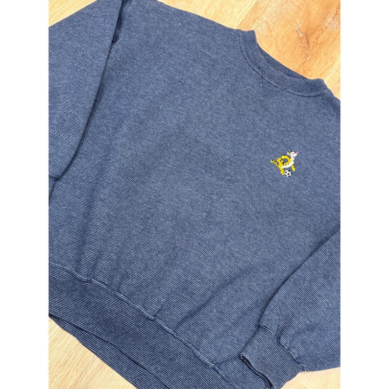 May include: A blue crew neck sweatshirt with a small embroidered design of a cartoon character playing soccer. The sweatshirt has a ribbed texture and ribbed cuffs and hem. The fabric appears to be a medium weight.