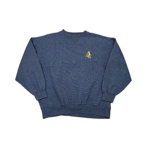 May include: A blue crew neck sweatshirt with a small embroidered design on the upper left chest. The design appears to be a gold emblem with stars. The sweatshirt has long sleeves and ribbed cuffs and hem.