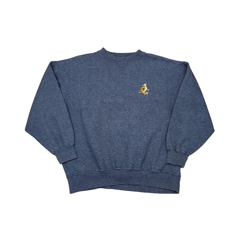 May include: A blue crew neck sweatshirt with a small embroidered design on the upper left chest. The design appears to be a gold emblem with stars. The sweatshirt has long sleeves and ribbed cuffs and hem.