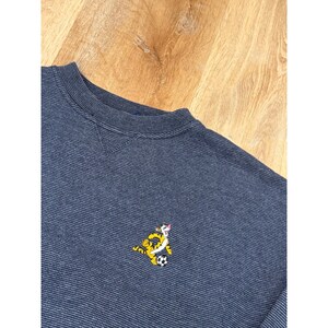 May include: A navy blue sweatshirt with a crew neck and a small embroidered cartoon tiger playing soccer. The sweatshirt has a textured pattern and is made of a soft material.