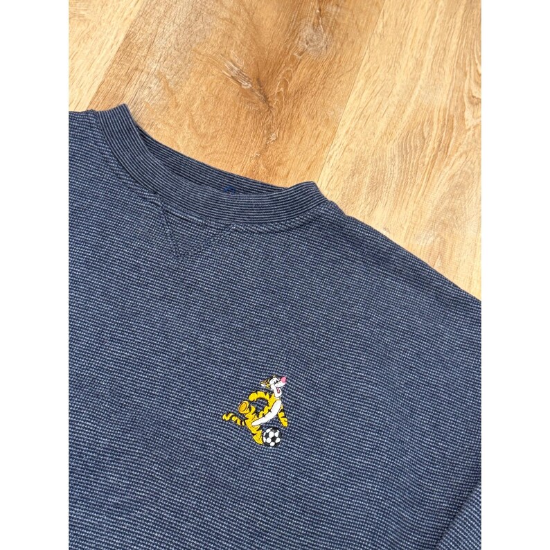 May include: A navy blue sweatshirt with a crew neck and a small embroidered cartoon tiger playing soccer. The sweatshirt has a textured pattern and is made of a soft material.