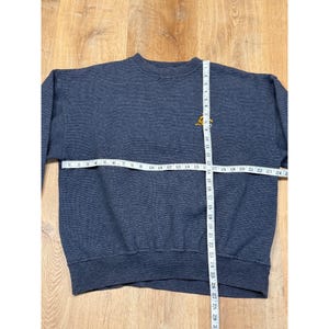 May include: A navy blue sweatshirt with a small yellow emblem on the upper chest. A measuring tape is laid across the chest, showing a width of approximately 24 inches. The sweatshirt is made of a textured fabric.