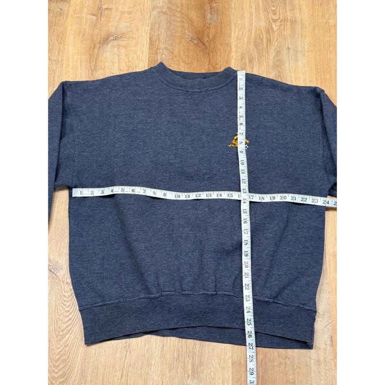 May include: A navy blue sweatshirt with a small yellow emblem on the upper chest. A measuring tape is laid across the chest, showing a width of approximately 24 inches. The sweatshirt is made of a textured fabric.