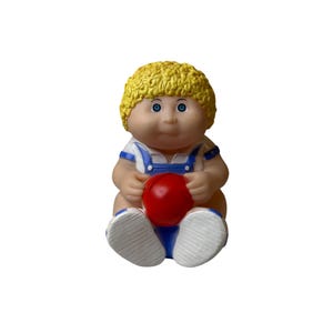 May include: A vintage Cabbage Patch Kid doll figurine. The doll has blonde, curly hair, blue eyes, and is wearing a blue and white outfit with a red ball. The doll is sitting down with its legs crossed.