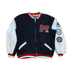 May include: A black and white baseball jacket with red and white trim. The jacket has a zipper closure and features a large red "M" with "World Tour" written on a blue banner. The sleeves have white fabric with various embroidered patches.