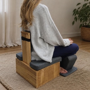 May include: A wooden meditation bench featuring a low-profile rectangular base with a vertical backrest support. The bench includes dark gray cushioned seating pads on the top and a matching gray floor mat, with a black strap attached to the backrest.