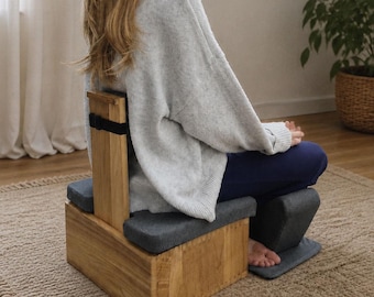 May include: A wooden meditation seat featuring a rectangular base with a vertical backrest and a black strap. The seat includes dark gray cushioned pads on the base and a separate matching gray cushioned support piece for the lower legs.