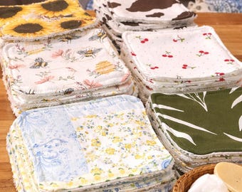 May include: A collection of square reusable face cloths featuring various patterns including sunflowers, bees, cow print, cherries, floral designs, and solid olive green. The text reads NEW! REUSABLE Face Cloths and ECO-FRIENDLY WASHABLE SO CUTE!