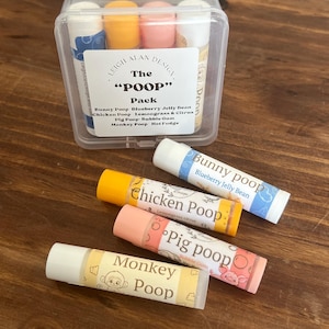 May include: A clear plastic case containing four lip balm tubes labeled The Poop Pack, featuring flavors Bunny Poop Blueberry Jelly Bean, Chicken Poop Lemongrass & Citrus, Pig Poop Bubble Gum, and Monkey Poop Hot Fudge, with four individual tubes displayed in front.