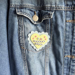 May include: A blue denim jacket featuring a heart-shaped felt patch pinned to the chest pocket. The patch has a light blue scalloped edge, a pale yellow center with small pink flower accents, and white letter beads spelling I HEART HOCKEY BOYS.