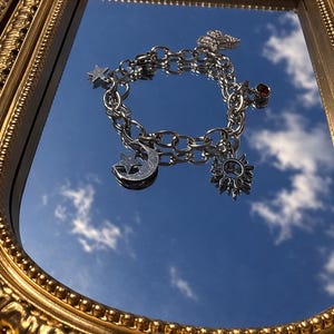 May include: A silver charm bracelet with celestial-themed charms, including a crescent moon, star, and sun. The bracelet also features a small red gemstone and a butterfly charm. The bracelet is displayed on a mirror with a gold frame.
