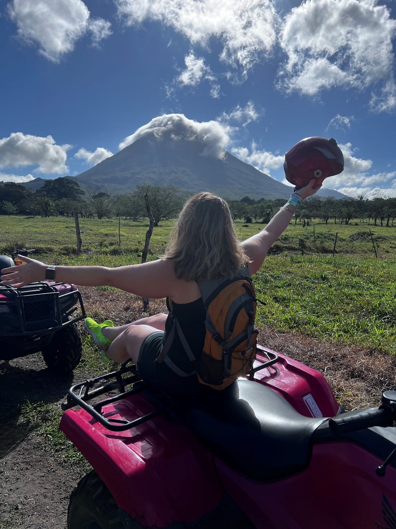 May include: A person wearing a black tank top and a yellow backpack sits on a pink ATV, holding a red helmet in one hand and looking up at a mountain in the distance.