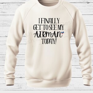 May include: A cream-colored sweatshirt with the text "I FINALLY GET TO SEE MY AIRMAN TODAY!" in black lettering. A small American flag heart is next to the word "AIRMAN".