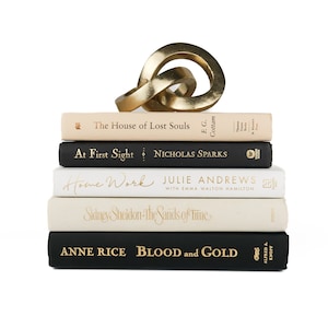 May include: A stack of five books with gold lettering on the spines. The books are arranged from top to bottom: The House of Lost Souls, At First Sight, Home Work, Sidney Sheldon: The Sands of Time, and Anne Rice: Blood and Gold. A gold metal sculpture of a knot sits on top of the stack.
