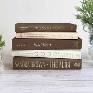 May include: A stack of five hardcover books with gold lettering. The books are in shades of brown and white. Titles include "The Secret Scripture," "Conversations with God," "Rebel Heart," "How to Know God," and "The Alibi."