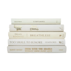 May include: A stack of five white books with gold lettering on the spines. The titles are "A Far Country", "Katherine Stone", "Happy Endings", "Dean Koontz | Breathless", "Too Small to Ignore", and "Swim With the Sharks Without Being Eaten Alive".