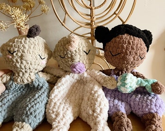 Puede incluir: Tres juguetes de peluche de crochet con diferentes tonos de piel. Los juguetes llevan atuendos de diferentes colores y tienen diferentes expresiones faciales. Los juguetes están sentados sobre una superficie de madera.