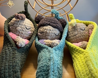 Puede incluir: Tres juguetes de peluche tejidos a crochet en forma de bebés durmiendo, cada uno acurrucado en una manta tejida a crochet en forma de vaina de guisante. Las mantas son verdes, azules y amarillas.