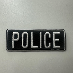 May include: Black rectangular patch with the word "POLICE" embroidered in white. The patch has a white border and is likely made of fabric. The text is centered on the patch.