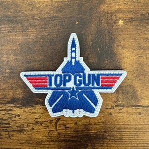 May include: An embroidered patch featuring a blue fighter jet silhouette with a central star and wings. The text TOP GUN is displayed in blue letters across the wings, which are accented with horizontal red stripes and a white border outline.